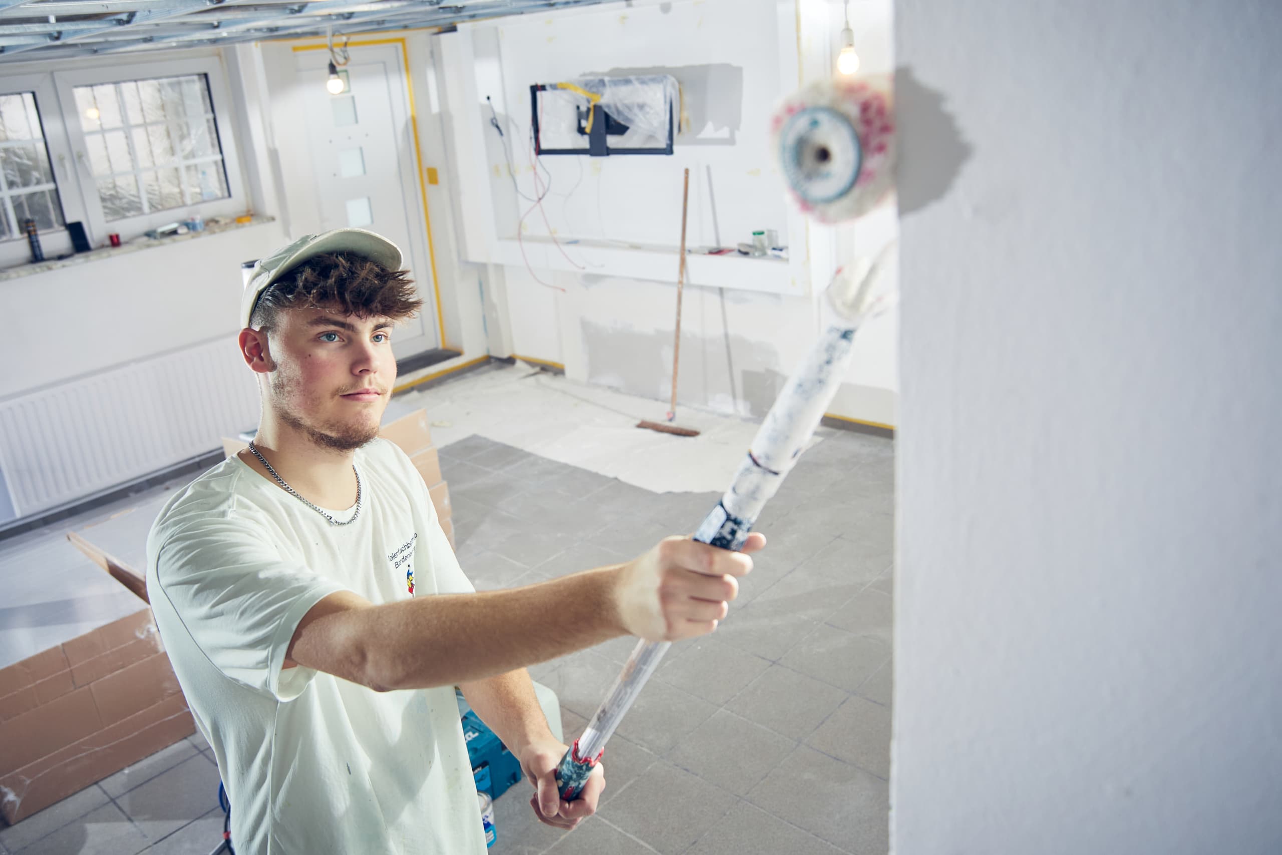 Junger Maler im weißen T-Shirt und Cap streicht mit einer Farbrolle eine Wand in einem Renovierungsraum